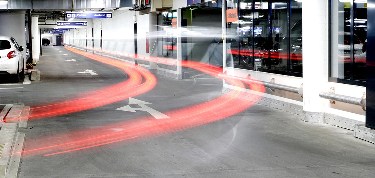 Weg in Richtung Ausfahrt in der Tiefgarage Stachus; durch Mehrfachbelichtung schweben Rücklichter eines Autos als rote Streifenspuren im Raum